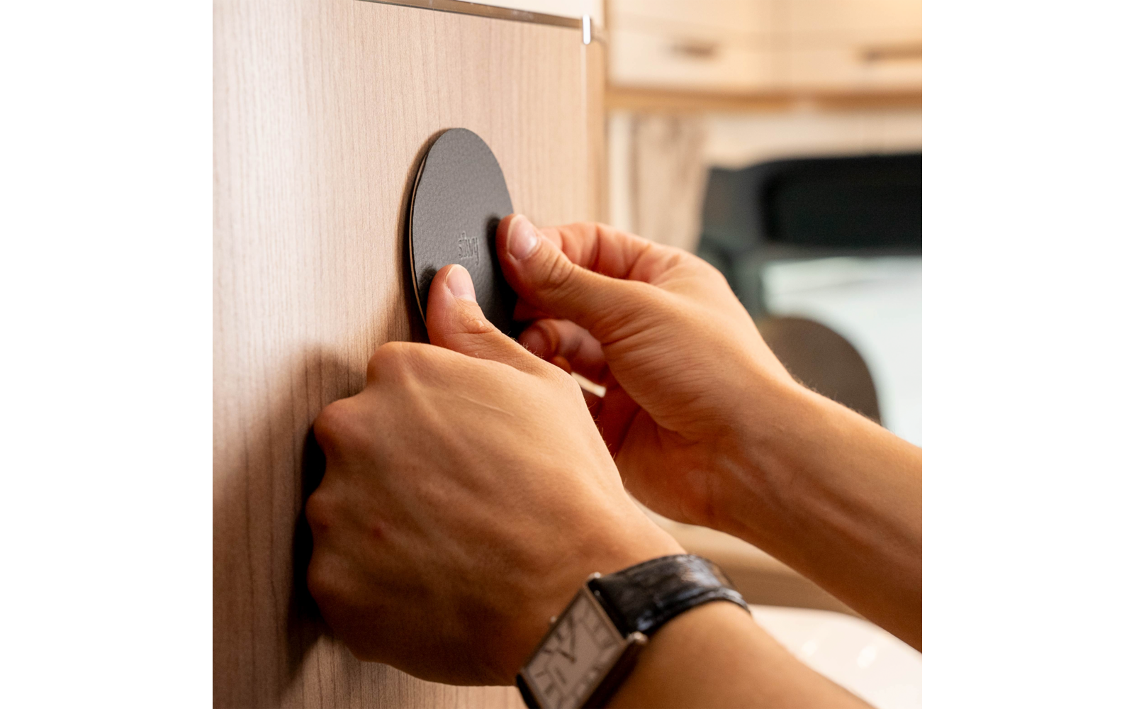 Zwei Hände halten und drücken eine runde schwarze Scheibe an eine Holzschranktür; ein Handgelenk trägt eine Armbanduhr; unscharfer Innenraum im Hintergrund