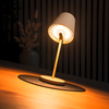 Weiße Tischlampe mit geneigtem Schirm und sichtbarer, leuchtender Birne auf rundem Untersetzer auf einem Holztisch; im Hintergrund vertikale Lamellen und links eine Vase mit getrockneten Blumen