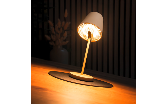 Weiße Tischlampe mit geneigtem Schirm und sichtbarer, leuchtender Birne auf rundem Untersetzer auf einem Holztisch; im Hintergrund vertikale Lamellen und links eine Vase mit getrockneten Blumen