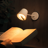 Hand hält ein aufgeschlagenes Buch unter einer eingeschalteten weißen Wandlampe; Holzvertäfelung im Hintergrund, Pflanzenblätter links