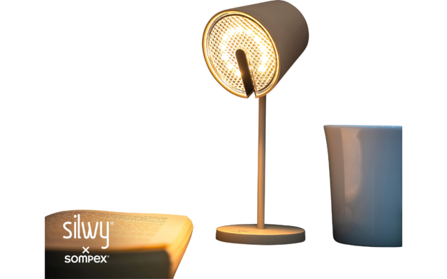 Lampe de table allumée, livre ouvert à côté, tasse sur un dessous de verre sombre, mur en bois en arrière-plan, logo de marque 'silvy x sompex' en bas à gauche