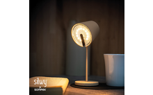 Eingeschaltete Tischlampe, daneben aufgeschlagenes Buch, Tasse auf dunklem Untersetzer, holzvertäfelte Wand im Hintergrund, Markenlogo 'silvy x sompex' unten links