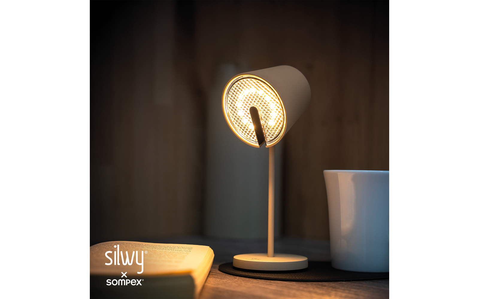 Eingeschaltete Tischlampe, daneben aufgeschlagenes Buch, Tasse auf dunklem Untersetzer, holzvertäfelte Wand im Hintergrund, Markenlogo 'silvy x sompex' unten links