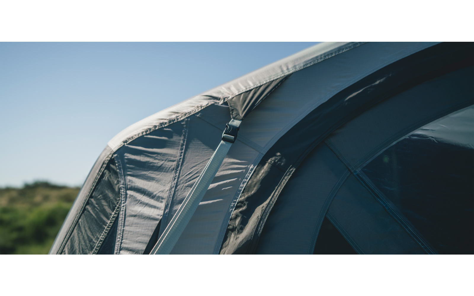 Nahaufnahme einer grau-blauen Zeltwand mit Riemen und Verschluss, blauem Himmel und Vegetation im Hintergrund