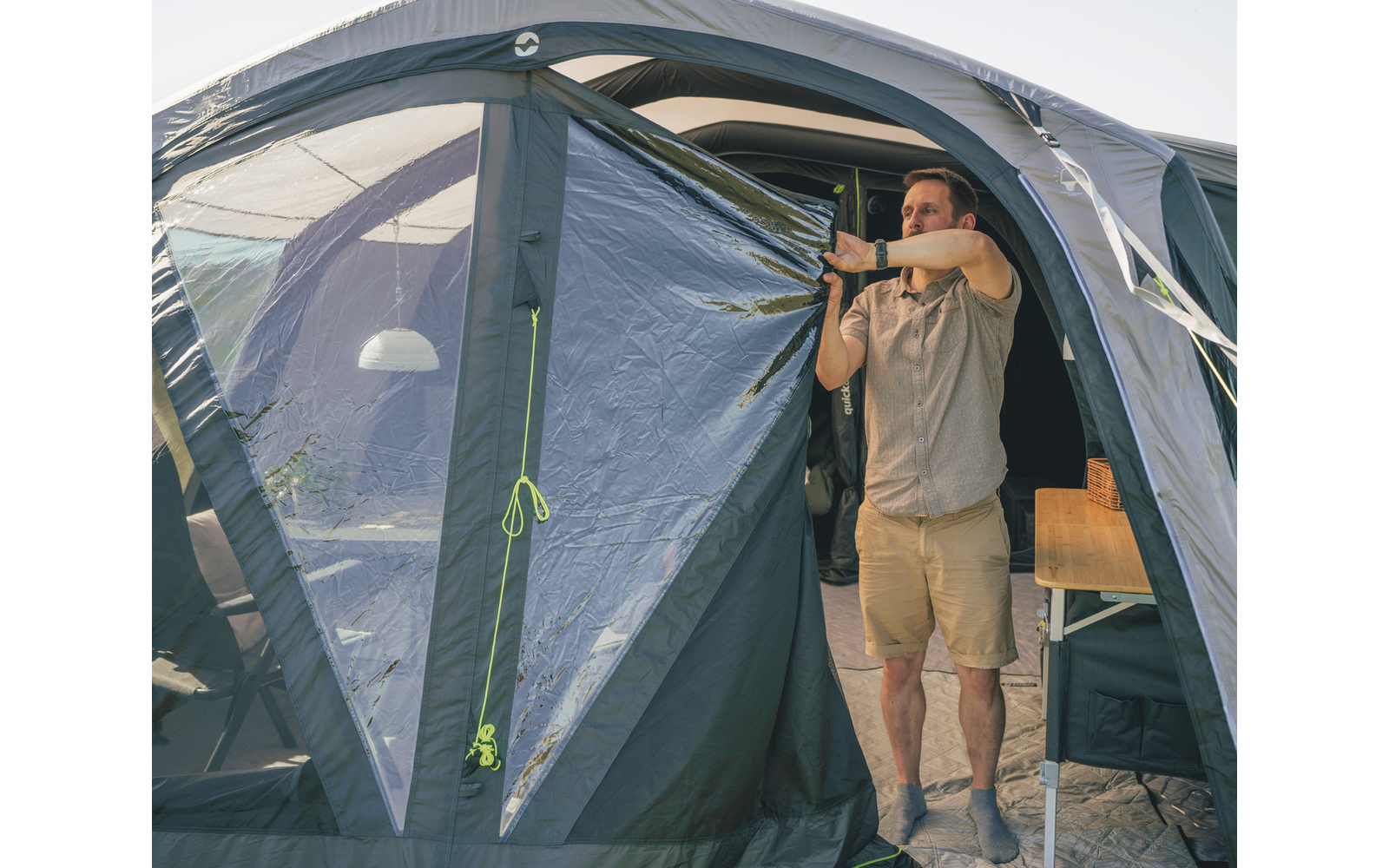 Mann steht in der geöffneten Eingangsklappe eines grauen Zelts und hält die Türflappe zur Seite; im Inneren sichtbar: kleiner Holztisch und Hängelampe