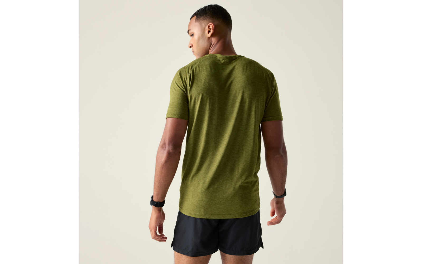 Personne aux cheveux courts et foncés, vue de dos, légèrement tournée vers la gauche, portant un T‑shirt vert olive et un short de sport noir court, bracelets aux deux poignets, arrière‑plan uni et clair