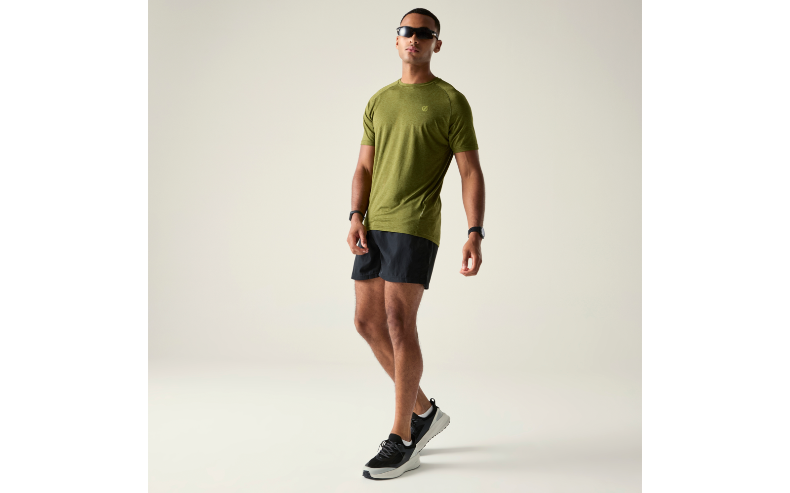 Personne portant des lunettes de soleil, un t-shirt vert olive, un short noir et des baskets noires sur fond clair