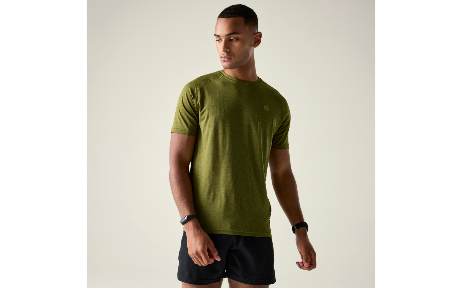 Personne debout en t‑shirt vert olive à manches courtes et short noir, regard tourné vers la gauche, fond uni clair, bracelets aux deux poignets