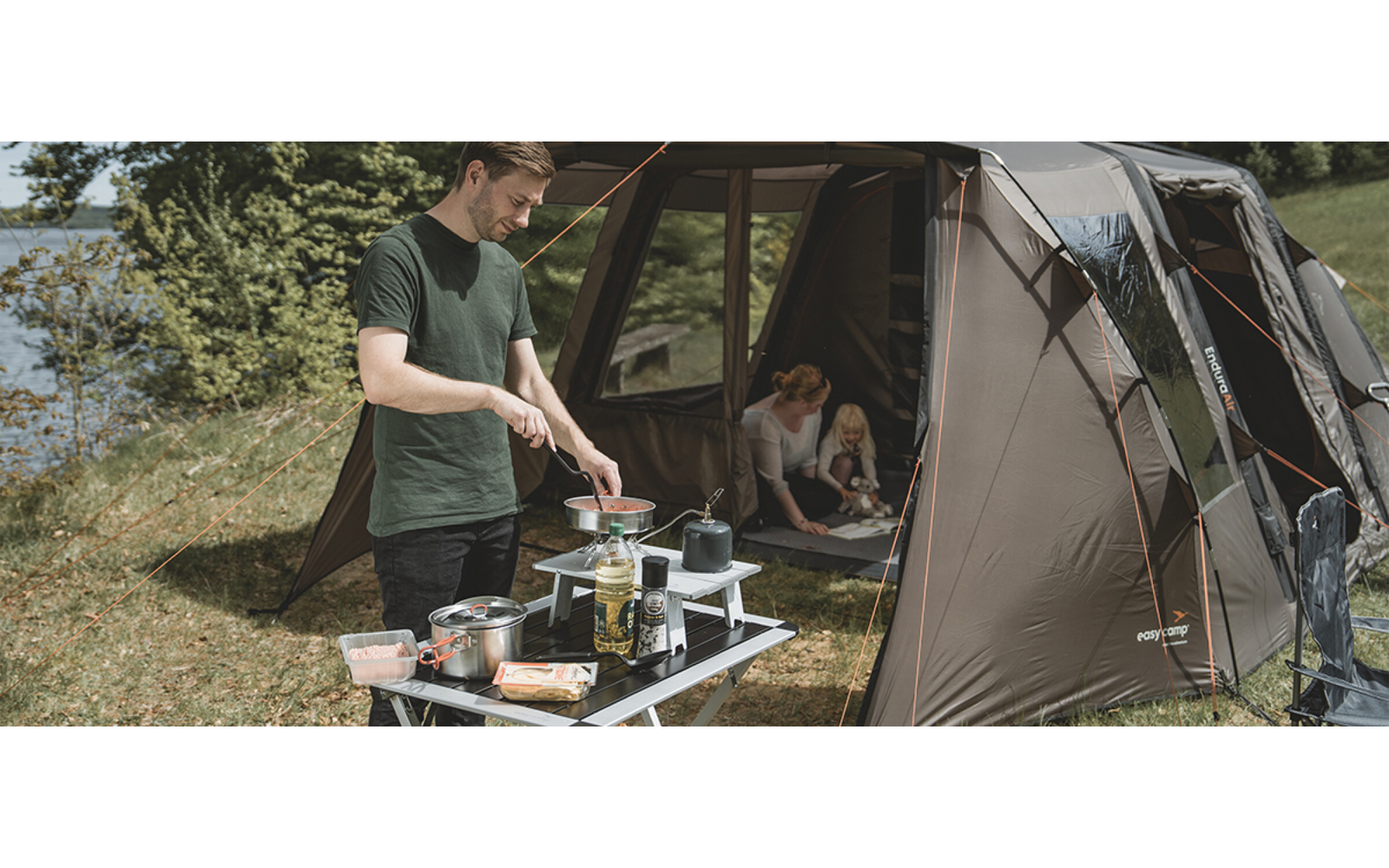 Mann rührt an einem Campingkocher auf einem Klapptisch; daneben Zelt mit Frau und Kind; See und Bäume im Hintergrund; Campingausrüstung auf dem Tisch