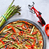 Tarte ronde dans un moule avec croûte, garnie d'asperges vertes, de lamelles de poivron rouge, de tranches violet foncé et d'herbes ; à gauche une botte d'asperges vertes, à droite un demi-poivron rouge et un petit modèle de vélo rouge