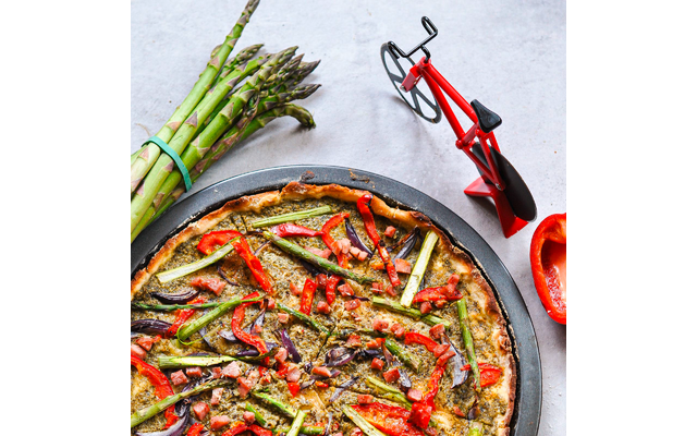 Tarte ronde dans un moule avec croûte, garnie d'asperges vertes, de lamelles de poivron rouge, de tranches violet foncé et d'herbes ; à gauche une botte d'asperges vertes, à droite un demi-poivron rouge et un petit modèle de vélo rouge