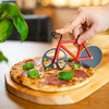 Main tenant un coupe-pizza rouge en forme de vélo au‑dessus d'une pizza aux tranches de salami et basilic sur une planche en bois