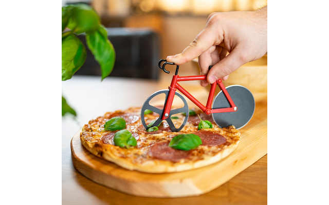 Main tenant un coupe-pizza rouge en forme de vélo au‑dessus d'une pizza aux tranches de salami et basilic sur une planche en bois
