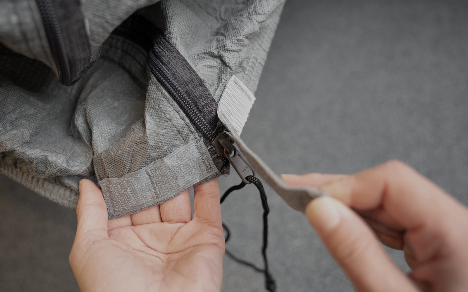Dos manos sostienen una tela gris texturizada y tiran de una cremallera con tirador
