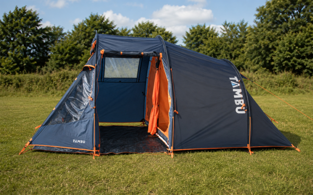 Tente de camping bleu foncé avec bordure orange et entrée avant ouverte, posée sur de l'herbe avec des arbres et un ciel partiellement nuageux en arrière‑plan, inscription 'TAMBU' visible sur le côté