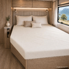 Tweepersoonsbed met beige hoeslaken, drie kussens, bekleed hoofdeinde, nachtkastje met vaas en groen, wandlampen, raam met uitzicht op meer en bergen, lichte houten inrichting