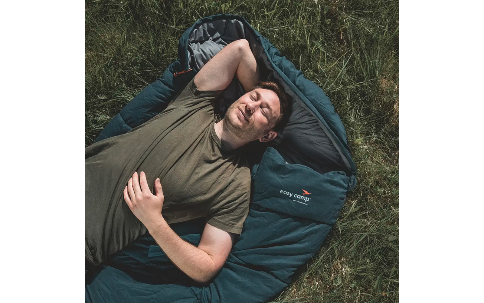 Mann liegt im Schlafsack auf Gras und lächelt