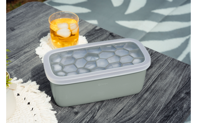 Rechteckiger hellgrüner Eiswürfelbehälter mit durchsichtigem Deckel und sechseckigen Mulden auf dunklem Holztisch; daneben Glas mit gelbem Getränk und Eis auf weißer gehäkelter Unterlage; Hintergrund mit Blattmuster