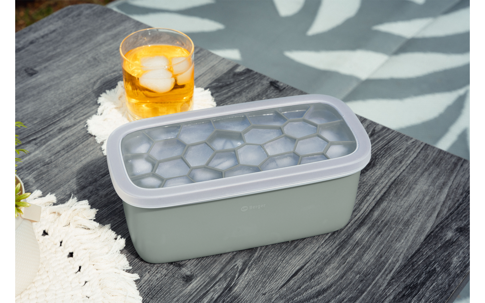 Rechteckiger hellgrüner Eiswürfelbehälter mit durchsichtigem Deckel und sechseckigen Mulden auf dunklem Holztisch; daneben Glas mit gelbem Getränk und Eis auf weißer gehäkelter Unterlage; Hintergrund mit Blattmuster