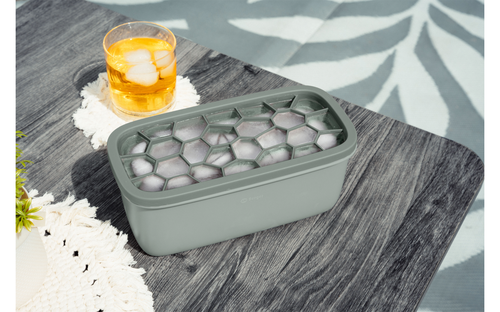 Grüner rechteckiger Behälter mit sechseckiger Wabenstruktur, gefüllt mit Eiswürfeln, auf dunklem Holztisch; daneben Glas mit bernsteinfarbener Flüssigkeit und Eis, weiße gehäkelte Unterlage und Blattmuster im Hintergrund