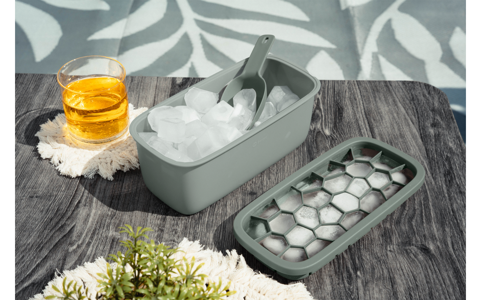Grauer Eisbehälter mit Eislöffel und Eiswürfeln, daneben sechseckiges Eiswürfelfach, Glas mit bernsteinfarbener Flüssigkeit auf weißer Unterlage, kleine grüne Pflanze und weißes Tuch auf dunklem Holztisch, Hintergrund mit Blattmuster