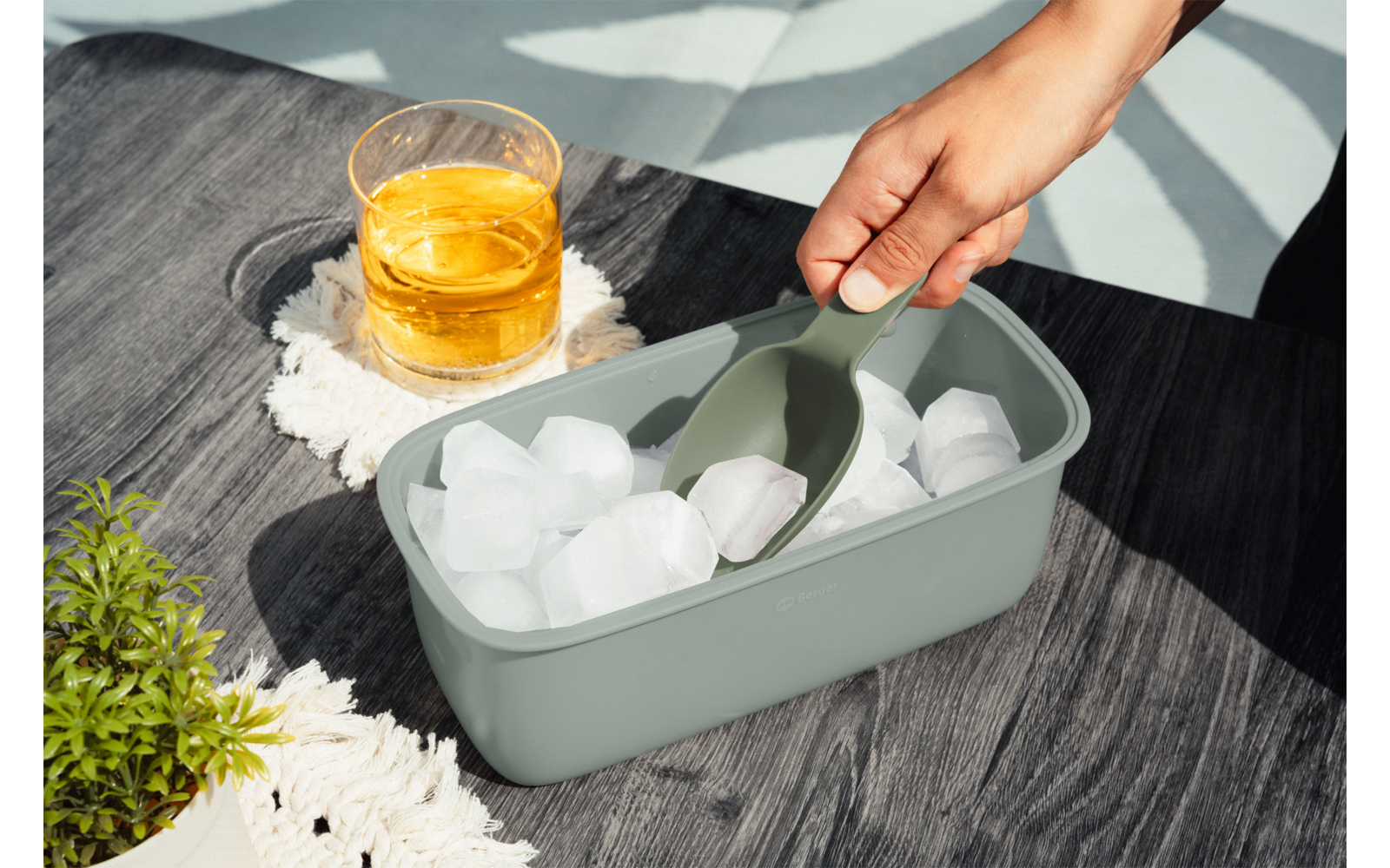 Hand mit Schaufel, die Eiswürfel aus rechtemckigem graugrünem Behälter auf dunklem Holztisch schöpft; daneben Glas mit Getränk auf gehäkeltem Untersetzer und kleine Topfpflanze