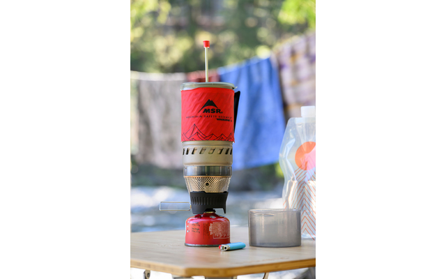 Fornellino a gas portatile rosso su una bomboletta, con una tazza di metallo, un accendino blu e una busta di plastica trasparente su un tavolo di legno; bucato sfocato sullo sfondo