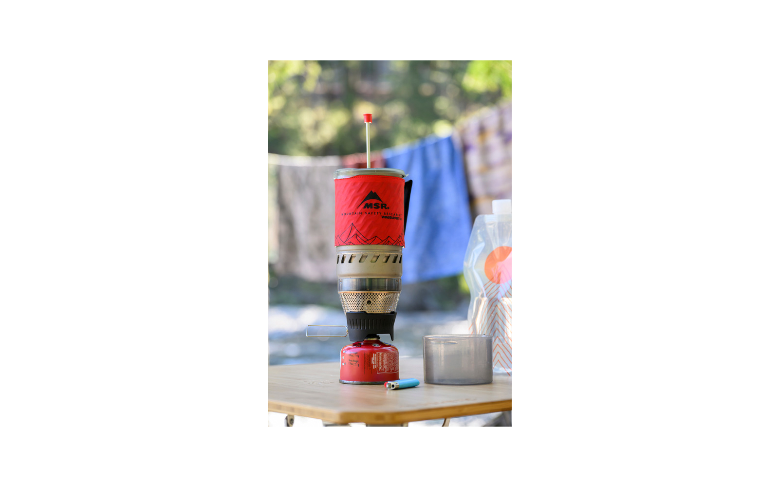 Fornellino a gas portatile rosso su una bomboletta, con una tazza di metallo, un accendino blu e una busta di plastica trasparente su un tavolo di legno; bucato sfocato sullo sfondo