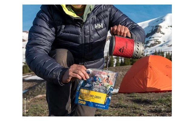 Persona con giacca imbottita blu versa acqua da una tazza rossa in una busta con la scritta 'Biscuits and Gravy'. Sullo sfondo una tenda arancione e montagne innevate