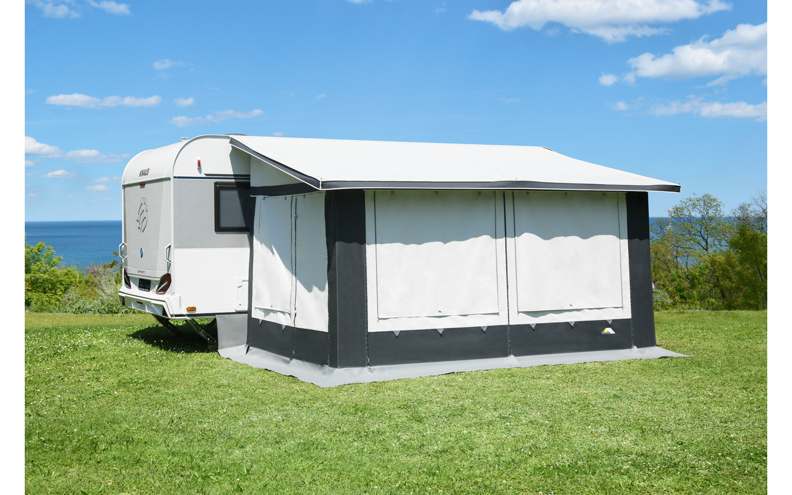 Weißer Wohnwagen mit angebautem Vorzelt auf grüner Wiese, im Hintergrund Meer und blauer Himmel mit wenigen Wolken