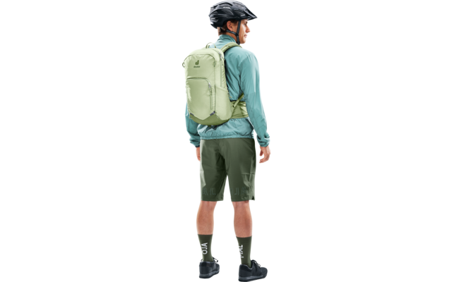 Seitenansicht einer Person mit Fahrradhelm, hellgrünem Rucksack, türkiser Jacke, dunkelgrünen Shorts, grünen Socken und schwarzen Schuhen, nach rechts blickend