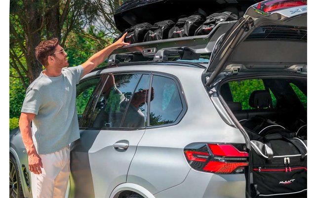 Ein Mann steht neben einem silbernen SUV; Heckklappe und Dachkoffer sind geöffnet, er greift in den Dachkoffer. Im Dachkoffer mehrere Helme, im Kofferraum eine schwarze Tasche; Bäume im Hintergrund