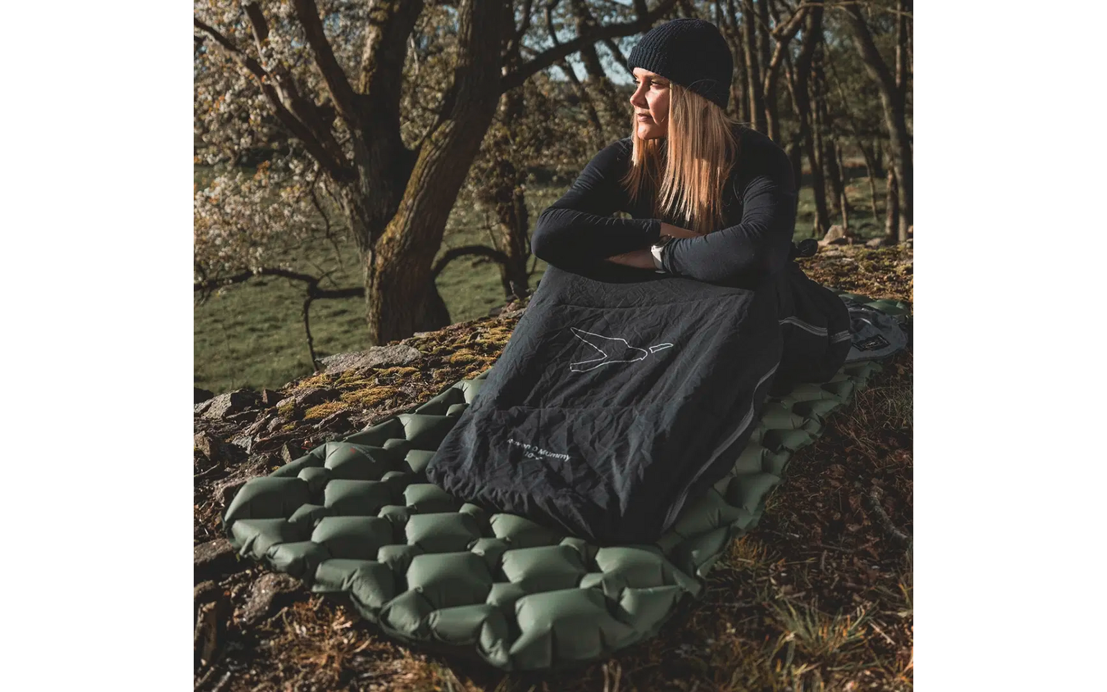 Frau sitzt draußen auf einer aufblasbaren Matte unter Bäumen, sie trägt eine Mütze und eine Outdoor-Jacke