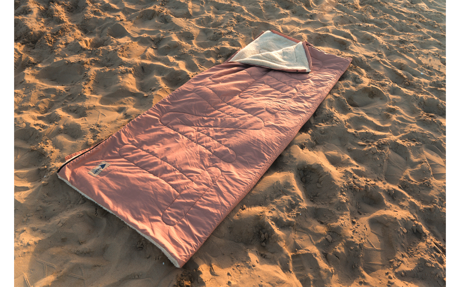 Sac de couchage rectangulaire orange avec doublure intérieure blanche visible, posé sur du sable ; aucune personne visible