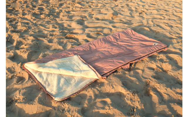 Sac de couchage rose ouvert avec doublure blanche posé sur du sable avec empreintes de pas et ondulations