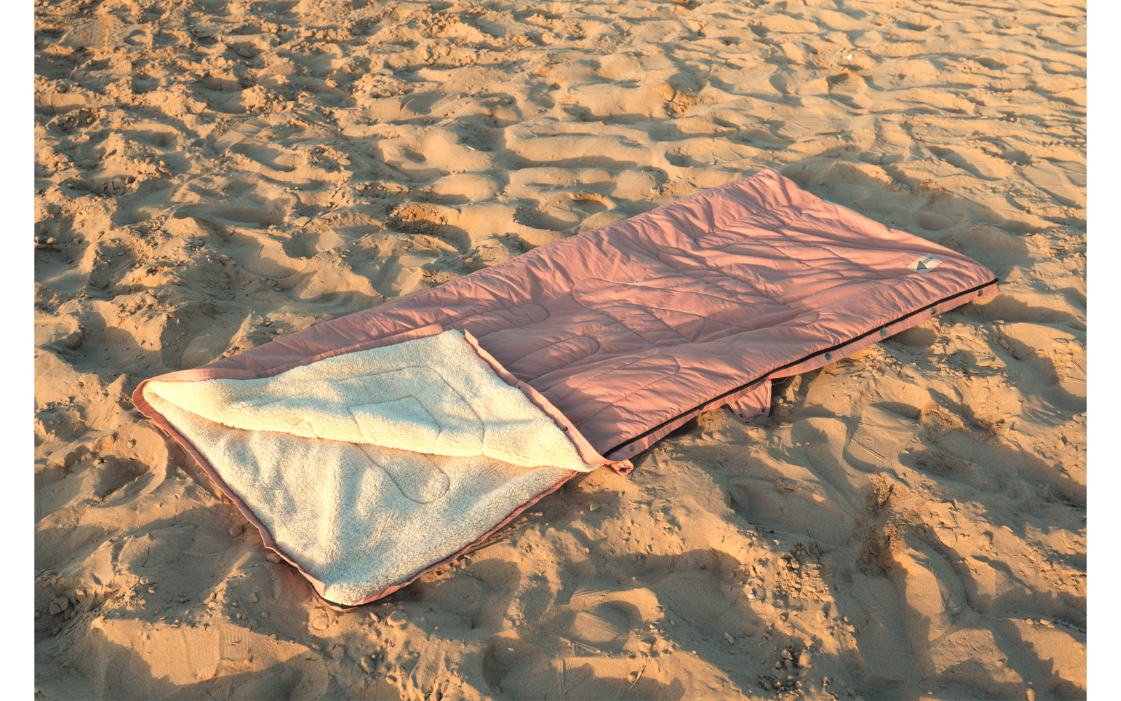 Sac de couchage rose ouvert avec doublure blanche posé sur du sable avec empreintes de pas et ondulations