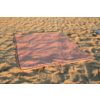 Couverture rectangulaire rose avec bord sombre posée sur du sable, plis sur la couverture et empreintes de pas dans le sable