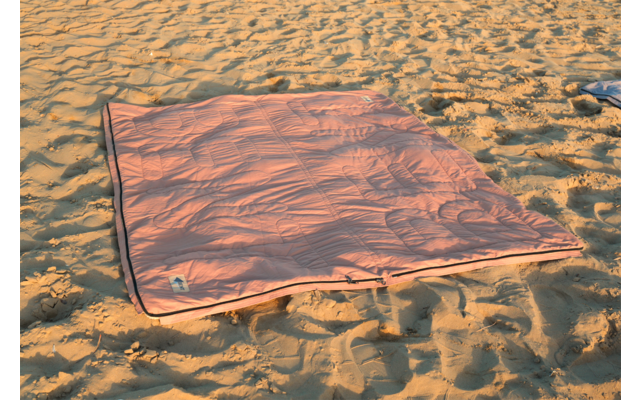 Couverture rectangulaire rose avec bord sombre posée sur du sable, plis sur la couverture et empreintes de pas dans le sable