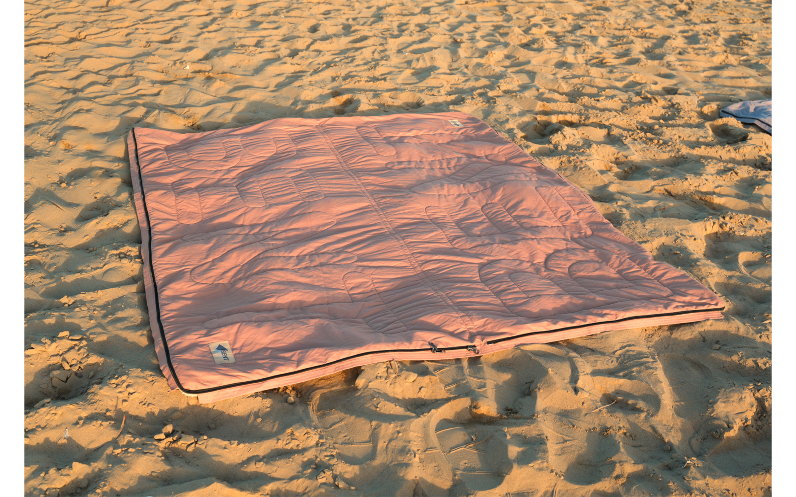Couverture rectangulaire rose avec bord sombre posée sur du sable, plis sur la couverture et empreintes de pas dans le sable