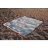Couverture blanche rectangulaire avec bord coloré posée sur du sable ; sable avec traces et ombres