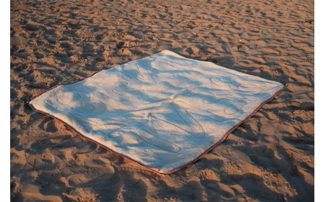 Couverture blanche rectangulaire avec bord coloré posée sur du sable ; sable avec traces et ombres