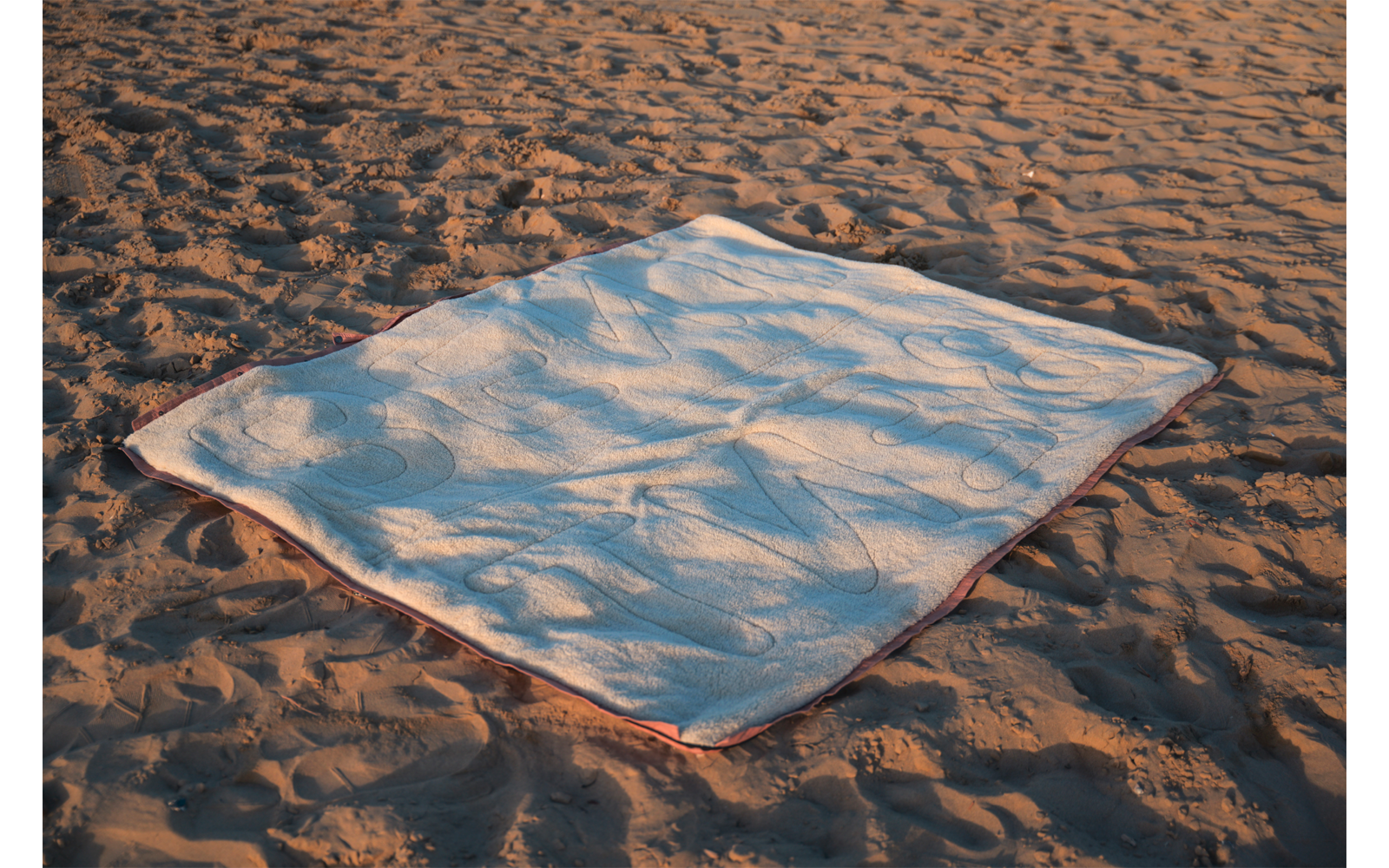 Couverture blanche rectangulaire avec bord coloré posée sur du sable ; sable avec traces et ombres