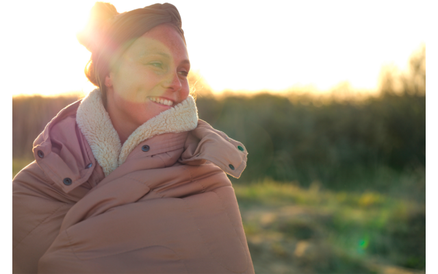 Personne portant un manteau matelassé épais et une coiffe, souriante, en contre-jour avec éblouissement du soleil, champ herbeux flou en arrière-plan