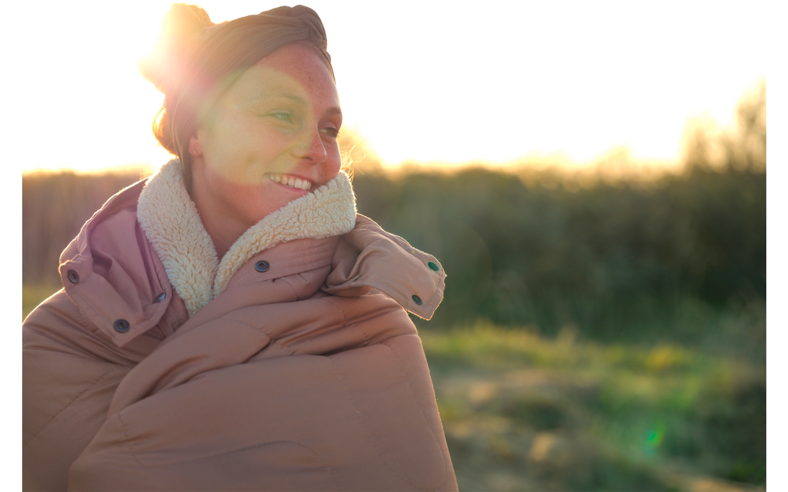 Personne portant un manteau matelassé épais et une coiffe, souriante, en contre-jour avec éblouissement du soleil, champ herbeux flou en arrière-plan