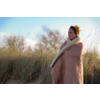 Femme avec les cheveux en chignon, enveloppée dans une couverture beige à doublure polaire, debout parmi l'herbe des dunes sur une dune de sable et regardant vers la droite