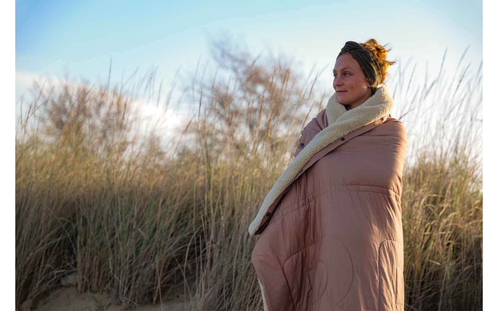 Femme avec les cheveux en chignon, enveloppée dans une couverture beige à doublure polaire, debout parmi l'herbe des dunes sur une dune de sable et regardant vers la droite