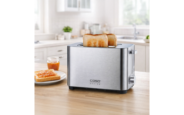 Edelstahl-Toaster mit zwei hochstehenden Toastscheiben auf der Arbeitsplatte; daneben Teller mit bestrichenem Toast und kleines Marmeladenglas; heller, unscharfer Küchenhintergrund