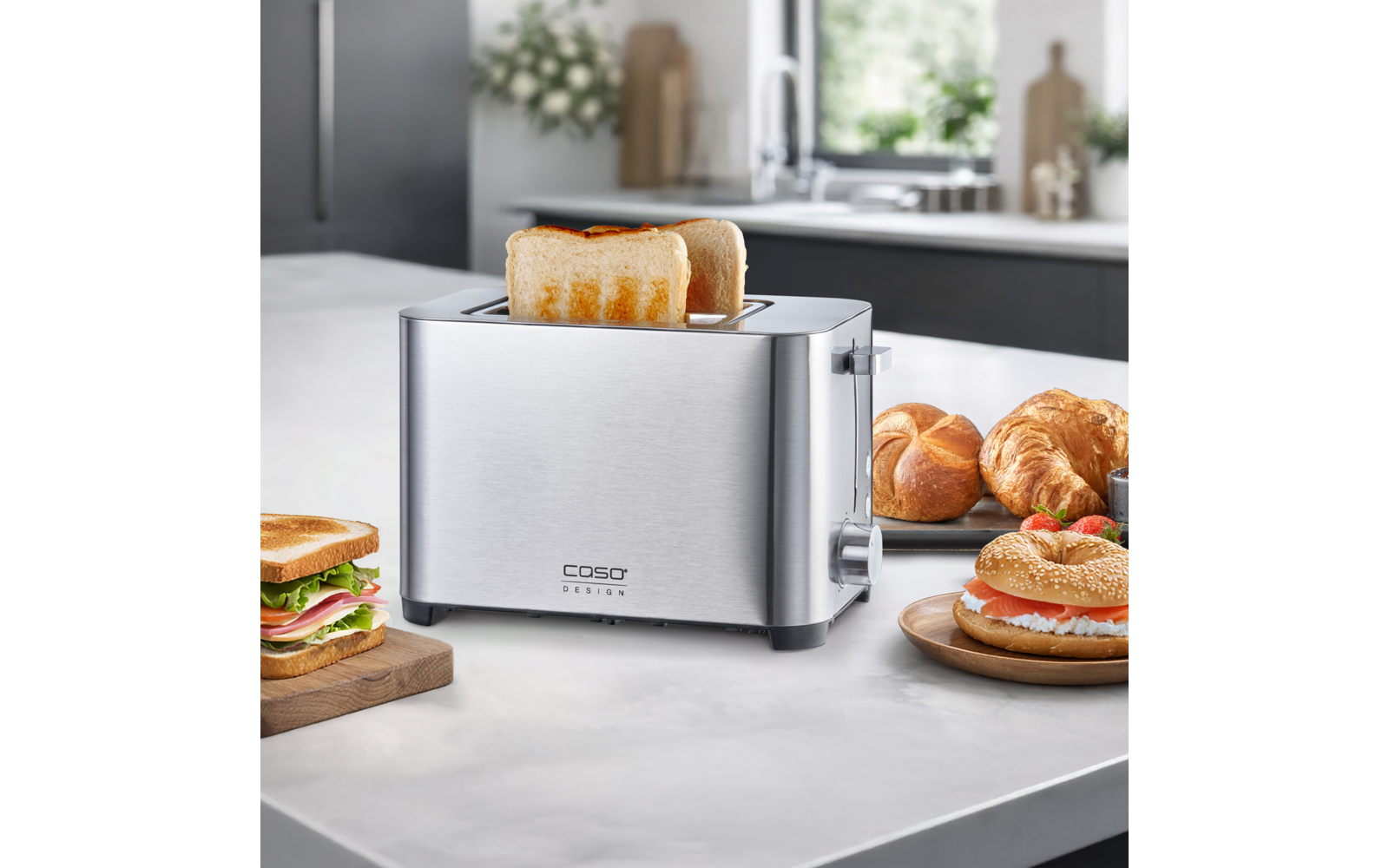 Edelstahl-Toaster mit zwei halb herausragenden Toastscheiben auf einer Küchenarbeitsplatte, daneben ein Sandwich, Croissants und ein Bagel