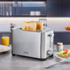 Edelstahl-Toaster mit zwei halb eingesteckten Toastscheiben auf einer Arbeitsplatte; daneben ein Glas Orangensaft, ein Teller mit Brot belegt mit Avocado und einer Tomatenscheibe sowie ein Ei im Eierbecher