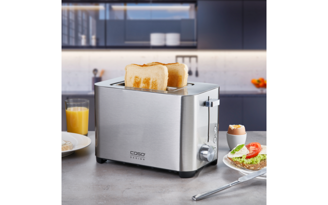 Edelstahl-Toaster mit zwei halb eingesteckten Toastscheiben auf einer Arbeitsplatte; daneben ein Glas Orangensaft, ein Teller mit Brot belegt mit Avocado und einer Tomatenscheibe sowie ein Ei im Eierbecher